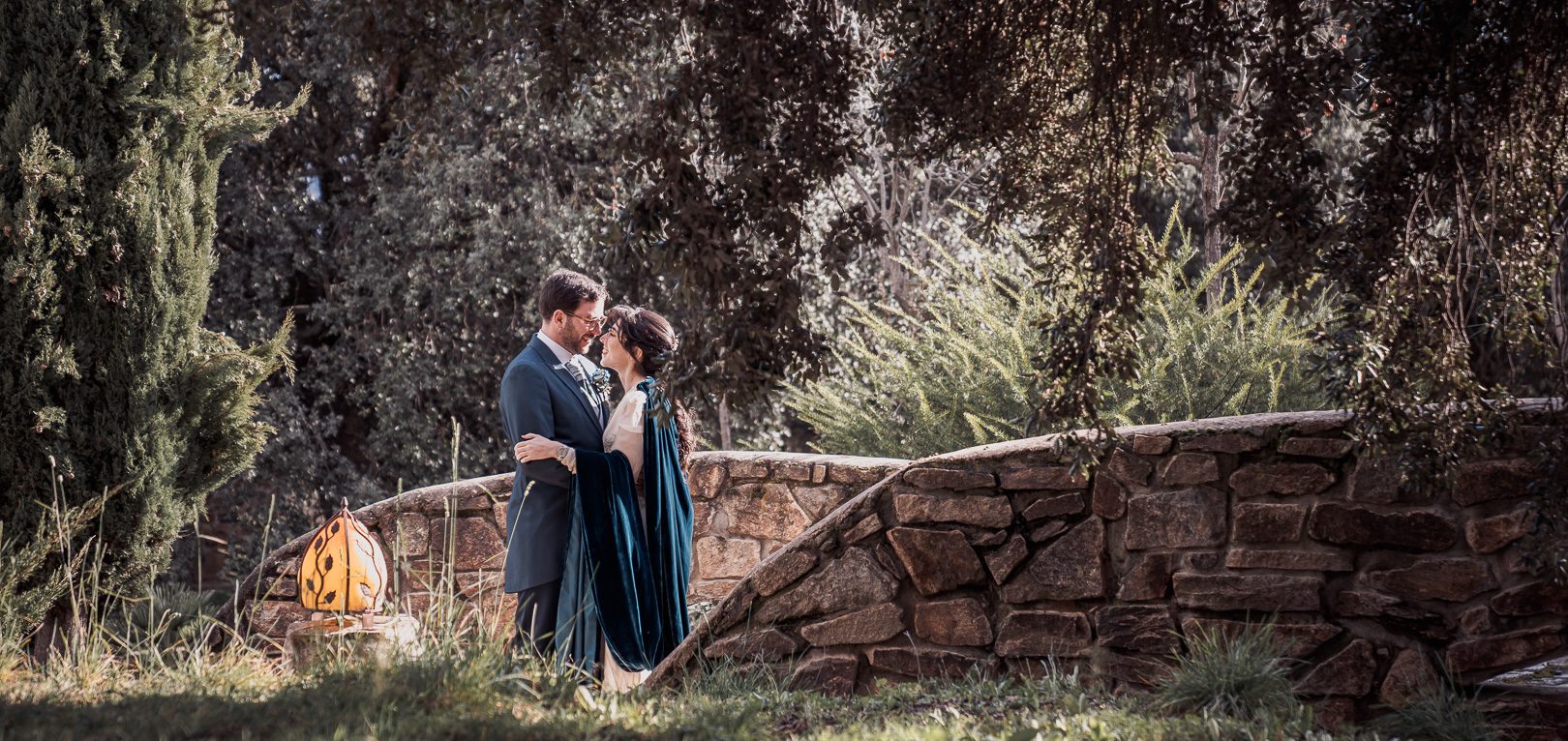 Pareja abrazándose en un entorno natural con vegetación y un muro de piedra.